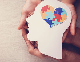 Hands holding artwork of a cutout head with a brain in the shape of a heart made with puzzle pieces.