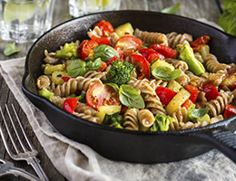 A cast iron pan filled with spiral pasta, tomatoes, broccoli and fresh herbs. 