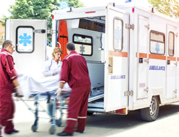 Paramedics in maroon scrubs rush a gurney out of an ambulance as a doctor in a white coat examines the patient.