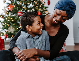 A smiling woman in a headscarf holds a smiling boy on her lap in front of a Christmas tree.