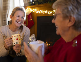 A woman laughs with an older woman as they hold mugs in front of a fireplace.