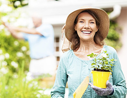 Older woman outdoors, wearing a sun hat and gloves and carrying a flowerpot and garden trowel.