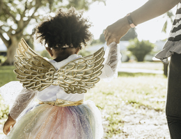 A little girl wearing a tutu and wings holds an adult’s hand.