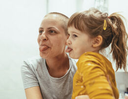A mother with a shaved head and daughter with a ponytail stick their tongues out.  