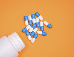 Blue and white capsules spill out of a white bottle on an orange background. 